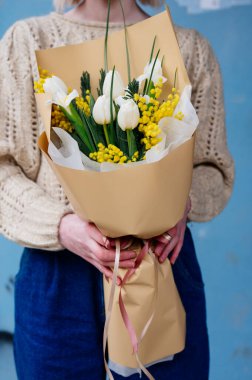 Beyaz lale buketi, narsis ve bej pakette mimoza. Mavi arka planda örülü kazak giyen çiçekçi kızın ellerinde..