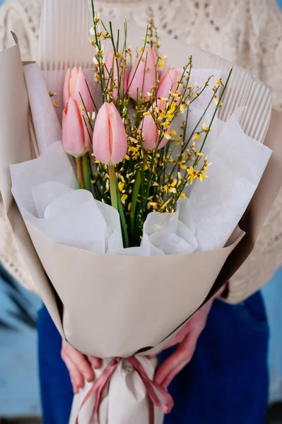 Bej pakette pembe lale buketi mavi arka planda örülü kazaklı çiçekçi kızın ellerinde..