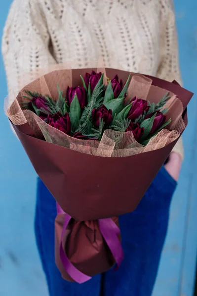 Kırmızı lale buketi ve mimoza yaprakları mavi arka planda örülü kazak giyen çiçekçi kızın ellerinde..