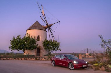 Murcia, İspanya - 26 Ekim 2019: Kırmızı bir Tesla Modeli ağaçlarla çevrili bir yel değirmeninin önüne park edilmiştir. Işıkları açık çünkü hava kararıyor..
