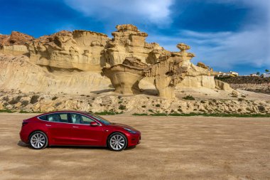 Bolnuevo, Murcia, İspanya - 7 Şubat 2020: Akdeniz kıyısında kaya oluşumlu kırmızı Tesla Model 3 elektrikli arabanın panoramik görüntüsü ve Bolnuevo, Murcia, İspanya 'da mavi gökyüzü.