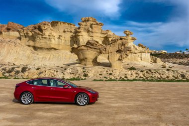 Bolnuevo, Murcia, Spain - February 7, 2020: panoramic view of a red Tesla Model 3 electric car with rock formation on the mediterranean coast and blue sky in Bolnuevo, Murcia, Spain. clipart