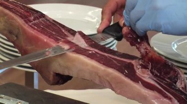 İberya jambonu kesen bir adamın ayrıntıları. Sağ el uzun, keskin bir bıçak kullanır ve sol el mavi lateks eldiven giyer ve jambon parçasını tutar..