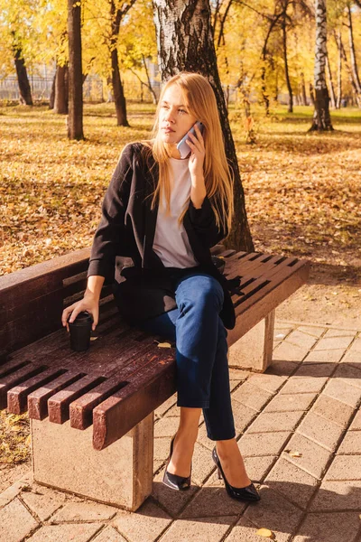 Young businesswoman talk on the phone in bench. Autumn park, sunny day.