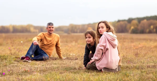 Happy dad and adult daughters sitting on the grass in a field at sunset. Happy man and little daughters spend time together. They\'re out for a walk. The family concept.
