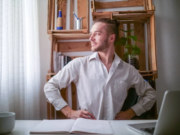 Beyaz gömlekli genç beyaz adam bir masada oturmuş gururlu ve tatmin olmuş bir şekilde pencereye bakıyor.