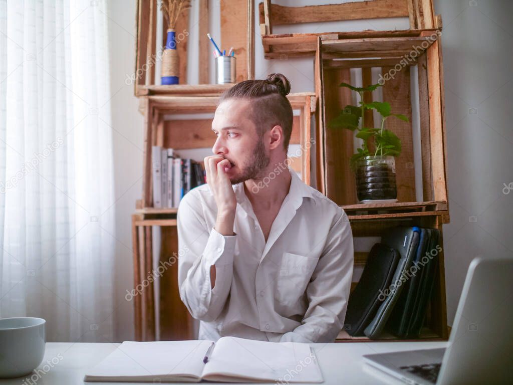 Young Caucasian Man in White Shirt Sitting at a Desk is Nervously Biting his Nails