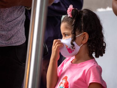 Medellin, Antioquia, Kolombiya - 3 Nisan 2021: Otobüs durağında Pembe Giyinmiş Küçük İspanyol Kız COVID Yüz Maskesiyle Bakıyor