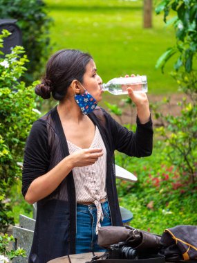 Yüz maskesi takan Latin Kadın, halk parkında plastik şişeden su içiyor.