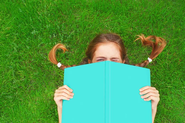 Küçük kız açık bir kitabın arkasından bakıyor, bir gözünü kapatıyor, yeşil çimlerin üzerinde yatıyor.