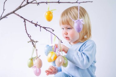 Sarışın çocuk arka planda paskalya dekoratif yumurtalarıyla ağaç dallarını süslüyor.