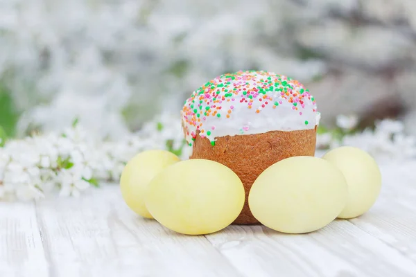 Bahçedeki beyaz ahşap bir masanın üzerinde renkli yumurtalarla süslü Paskalya pastası çiçek açan ağaçların arka planında