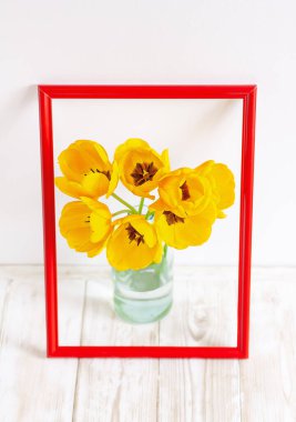 Sarı lale buketi cam vazoda, kırmızı çerçeveli beyaz bir duvara karşı.
