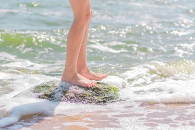 Genç bir kızın çıplak ayakları deniz kıyısındaki bir taşın üzerinde duruyor. Yaz tatili ve seyahat konsepti