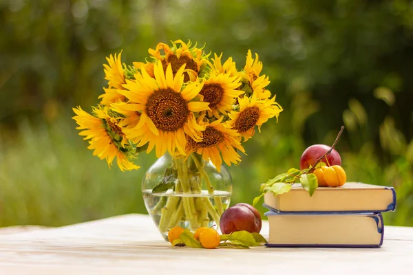 a stack of books and plums next to a glass vase with yellow sunflowers on an old wooden table in the garden