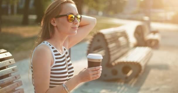 Vrouw die koffie drinkt in het park op de bank — Stockvideo