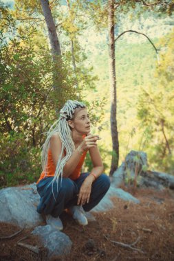 Rastafaryan bir kız Rastafaryan saçlarıyla bir taşın üzerinde oturur ve sigara içer.. 