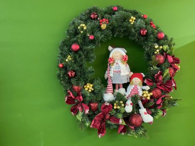 Yeşil duvarın sağ tarafında içinde bez bebekler olan bir Noel ağacı çelengi asılı..