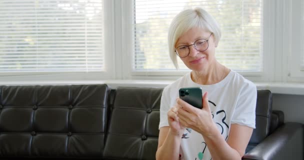 電話で熟女さん ソーシャルメディアアプリを使ってモバイルゲームをしている携帯電話の画面を見て 自宅のソファに座っている中年の女性 高齢者と近代的な技術の概念 — ストック動画