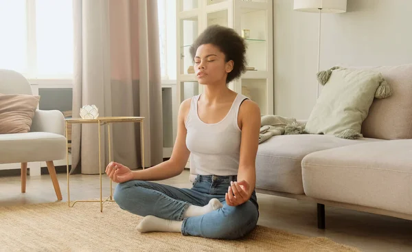 Afrikalı Amerikalı kadın meditasyon yapıyor ve evde oturuyor. Sakin kadın derin nefes alıyor ve gözleri kapalı dinleniyor. Stres ve huzur yok..