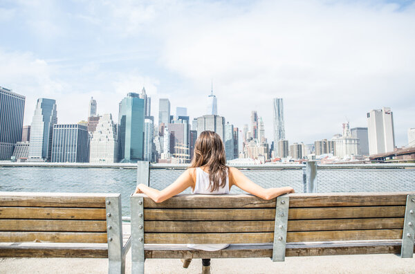 Woman sitting on a bench and looking at New York skyline - Tourist enjoys the view of Manhattan cityscape