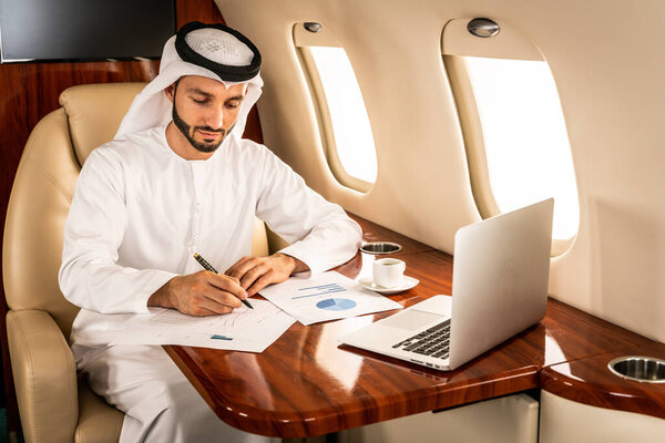 Arabic man wearing kandora in emirates style flying on exclusive private jet - Middle-eastern businessman with traditional dress flies in exclusive business class on airplane, concepts about business and trasportation