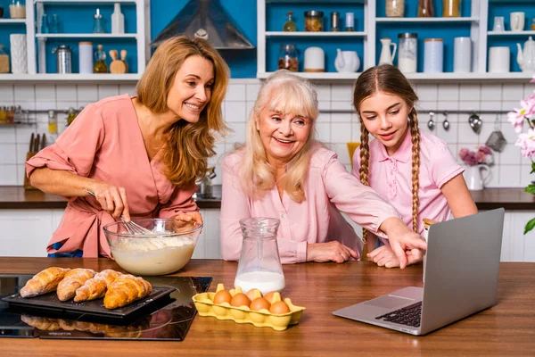 Beautiful senior woman and family baking in the kitchen - Grandmother preparing desserts at home with daughter and nephew looking at recipe online on computer laptop, concepts about cooking and healthy eating