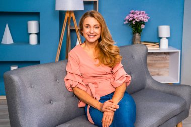 Güzel yetişkin kadın evde tek başına oturuyor ve kameraya bakıp gülümsüyor.