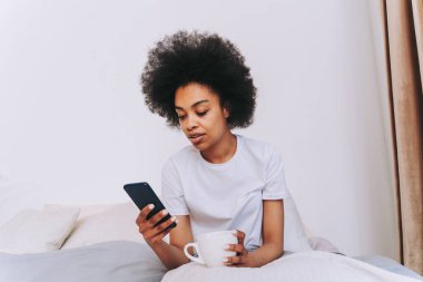 Afro-Amerikalı bir kız evde yatağında dinleniyor güzel bir kadın evde kahvaltı yapıyor ve akıllı telefonunu kontrol ediyor.