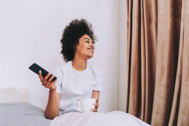 Afro-Amerikalı bir kız evde yatağında dinleniyor güzel bir kadın evde kahvaltı yapıyor ve akıllı telefonunu kontrol ediyor.