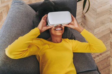 Evde VR kulaklıkla oynayan güzel bir kadın. Arttırılmış gerçeklik teknolojisiyle etkileşim halindeyken siyah bir kadın kanepeye uzanıyor.