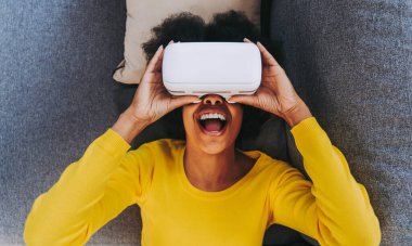 Evde VR kulaklıkla oynayan güzel bir kadın. Arttırılmış gerçeklik teknolojisiyle etkileşim halindeyken siyah bir kadın kanepeye uzanıyor.