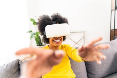 Evde VR kulaklıkla oynayan güzel bir kadın. Arttırılmış gerçeklik teknolojisiyle etkileşim halindeyken siyah bir kadın kanepeye uzanıyor.