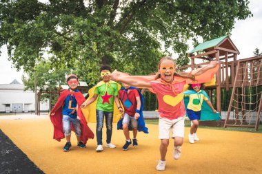 Multiracial group of young schooler wearing superhero costumes and having fun outdoors clipart