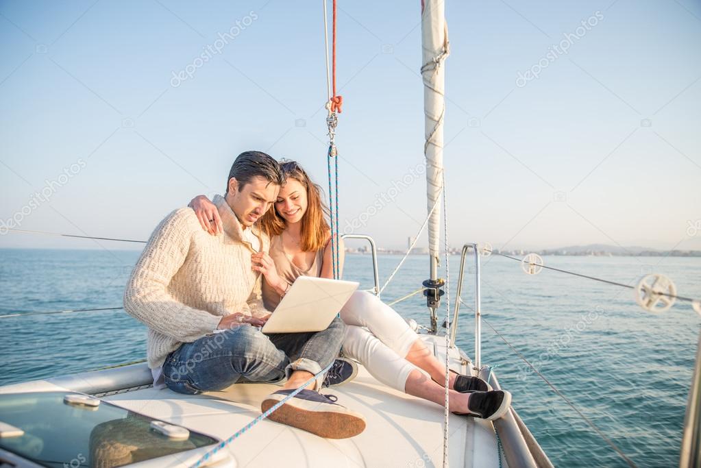 Couple sitting on a sailing boat looking at laptop computer - Beautiful woman and attractive man having fun on a boat while on vacation