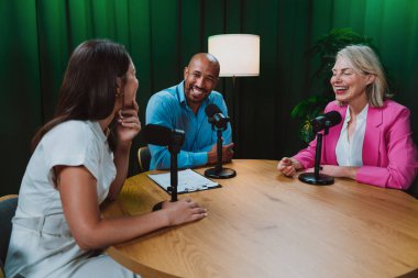 Üç farklı yetişkin sunucu, bir adam ve iki kadın, mikrofonlu bir masanın etrafında oturmuş, canlı bir podcast tartışmasına girişiyor ya da perdeli bir stüdyo ortamında röportaj yapıyorlar.