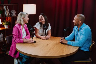 Profesyonel bir podcast stüdyosunda, mikrofonlar ve kameralarla yayın yapan, dinamik bir konuşma yapan üç kişiden oluşan bir grup.