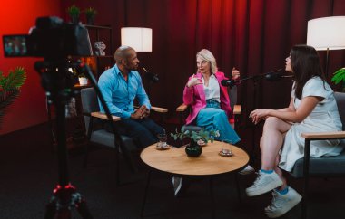 Profesyonel bir podcast stüdyosunda, mikrofonlar ve kameralarla yayın yapan, dinamik bir konuşma yapan üç kişiden oluşan bir grup.