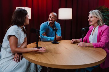 Bir grup insan bir masanın etrafında oturuyor, aktif olarak bir podcast kaydına bağlanıyor, bir kahkahayı paylaşıyor ve profesyonel bir stüdyo ortamında içerik yaratıyorlar.