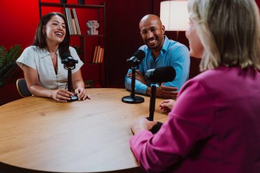 Bir grup insan bir masanın etrafında oturuyor, aktif olarak bir podcast kaydına bağlanıyor, bir kahkahayı paylaşıyor ve profesyonel bir stüdyo ortamında içerik yaratıyorlar.