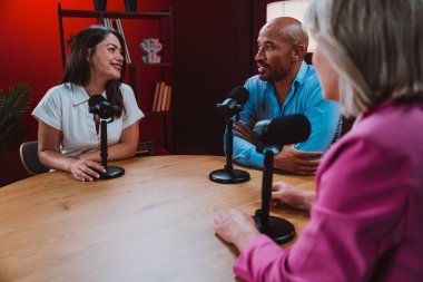 Bir grup insan bir masanın etrafında oturuyor, aktif olarak bir podcast kaydına bağlanıyor, bir kahkahayı paylaşıyor ve profesyonel bir stüdyo ortamında içerik yaratıyorlar.