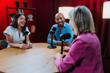 Bir grup insan bir masanın etrafında oturuyor, aktif olarak bir podcast kaydına bağlanıyor, bir kahkahayı paylaşıyor ve profesyonel bir stüdyo ortamında içerik yaratıyorlar.