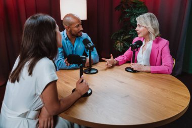 Bir grup insan bir masanın etrafında oturuyor, aktif olarak bir podcast kaydına bağlanıyor, bir kahkahayı paylaşıyor ve profesyonel bir stüdyo ortamında içerik yaratıyorlar.