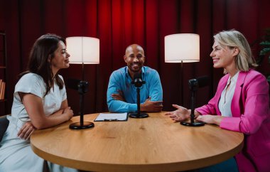 Üç farklı yetişkin sunucu, bir adam ve iki kadın, mikrofonlu bir masanın etrafında oturmuş, canlı bir podcast tartışmasına girişiyor ya da perdeli bir stüdyo ortamında röportaj yapıyorlar.