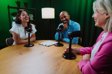 Üç farklı yetişkin sunucu, bir adam ve iki kadın, mikrofonlu bir masanın etrafında oturmuş, canlı bir podcast tartışmasına girişiyor ya da perdeli bir stüdyo ortamında röportaj yapıyorlar.