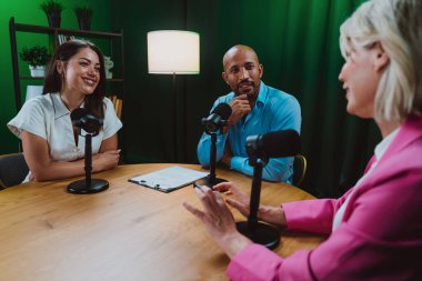 Üç farklı yetişkin sunucu, bir adam ve iki kadın, mikrofonlu bir masanın etrafında oturmuş, canlı bir podcast tartışmasına girişiyor ya da perdeli bir stüdyo ortamında röportaj yapıyorlar.