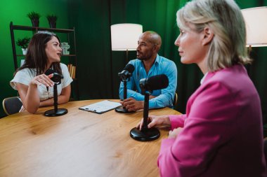 Üç farklı yetişkin sunucu, bir adam ve iki kadın, mikrofonlu bir masanın etrafında oturmuş, canlı bir podcast tartışmasına girişiyor ya da perdeli bir stüdyo ortamında röportaj yapıyorlar.