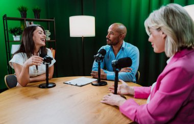 Üç farklı yetişkin sunucu, bir adam ve iki kadın, mikrofonlu bir masanın etrafında oturmuş, canlı bir podcast tartışmasına girişiyor ya da perdeli bir stüdyo ortamında röportaj yapıyorlar.