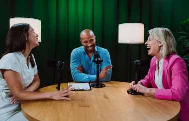 Üç farklı yetişkin sunucu, bir adam ve iki kadın, mikrofonlu bir masanın etrafında oturmuş, canlı bir podcast tartışmasına girişiyor ya da perdeli bir stüdyo ortamında röportaj yapıyorlar.