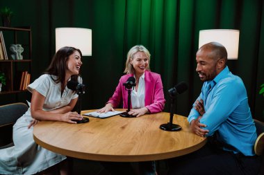 Üç farklı yetişkin sunucu, bir adam ve iki kadın, mikrofonlu bir masanın etrafında oturmuş, canlı bir podcast tartışmasına girişiyor ya da perdeli bir stüdyo ortamında röportaj yapıyorlar.